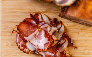 Two chubs of coppa and next to thinly sliced coppa on a cutting board and picture from above.