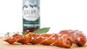 A closeup of il porcellino salumi's Breckenridge Brewery Avalanche Beer Smoked Cheddar Brats pictured from the side and cut into slices on a cutting board. A whole brat, herbs, spices, and a beer bottle are in the background.