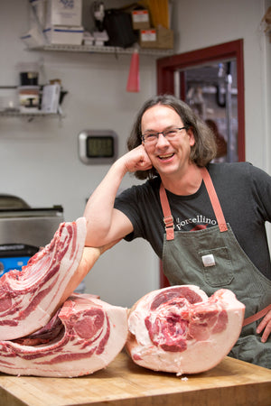 A picture of Bill Miner, who has long hair and is wearing a butchers apron, resting on a primal cut of a recently processed hog on a butcher table at his deli.
