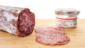 A chub of il porcellino salumi's large format Black Truffle Salami on the left side of a cutting board with thin slices in the foreground and a jar of black truffle salt to the right.