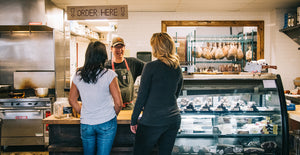Two women ordering from Bill Miner in il porcellino salumi's deli. The two women stand at the ordering counter next two the glass meat case with the drying room and stove in the background.