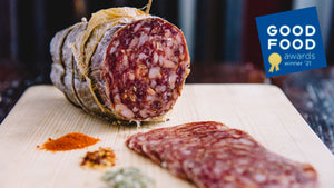 A large format chub of il porcellino salumi's Diablo Salami pictured head on with salami slices, herbs and spices in the foreground. The Good Food awards 2021 winner seal is on the image.