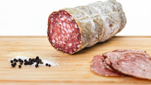A large format chub of il porcellino salumi's Genoa Salami on a cutting board with thin salami slices, salt, pepper, and garlic powder in the foreground.