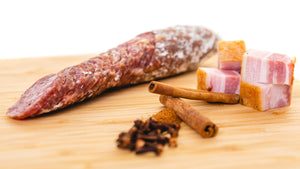 A chub of il porcellino salumi's Giffords Bacon Whiskey salami in a cutting board cut into slices. Chunks of bacon, cinnamon sticks and spices are in the foreground. 