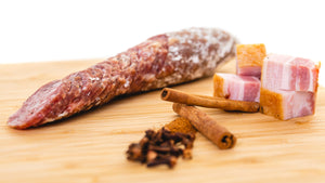 A chub of il porcellino salumi's Giffords Bacon Whiskey salami in a cutting board cut into slices. Chunks of bacon, cinnamon sticks and spices are in the foreground.