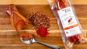 Two one-pound chubs of il porcellino salumi's 'Nduja Salami pictured from above on a cutting board. One chub has a chunk of salami removed with a spoon and the other is in packaging with herbs and spices between them.