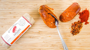 Two chubs of il porcellino salumi's 'Nduja salami pictured from above on a cutting board. One of the chubs has a spoon in it and the other is in packaging and herbs and spices are on the side.