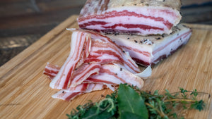 Slices and chubs of il porcellino salumi's Pancetta Tesa on a cutting board with green herbs in the foreground.
