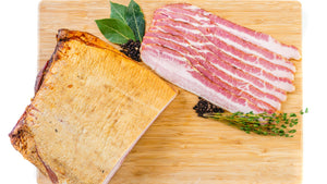 A slab and slices of il porcellino salumi's Peach Wood Smoked Bacon pictured from above on a cutting board with herbs and spices next to them.