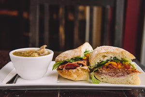 A cup of soup on a plate next to two halves of a sandwich and pictured head on.