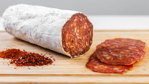 il porcellino salumi's large format Saucisson Basque salami picture up close on a cutting board with salami slices, herbs and spices in the foreground.