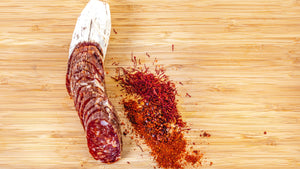 il porcellino salumi's Saucisson Basque salami pictured from above on a cutting board and cut into slices with herbs and spices next to it. 