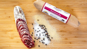 Two chubs of il porcellino salumi's Saucisson Sec salami pictured from above on a cutting board. One of the chubs is cut into slices and the other is in packaging with salt, peppercorns, and garlic powder between them.