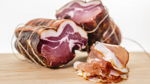 Slices of il porcellino salumi's Spanish Lomo on a cutting board with whole muscle chubs in the background.