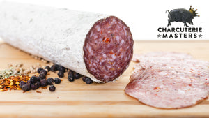 A chub of il porcellino salumi's large format Spiced Juniper salami pictured at an angle on a cutting board with salami slices, herbs and spices next to it. The charcuterie masters grand champion seal is placed on the picture.