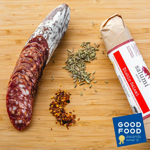 A picture from above of a spicy salami called Diablo salami in packaging and cut into slices on a cutting board with herbs and spices. Plus a Good Food Awards 2021 seal is placed on the image.