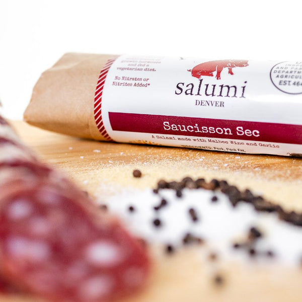 A close up of saucisson sec salami in packaging and on a cutting board with blurred salami slices, herbs and spices in the foreground.