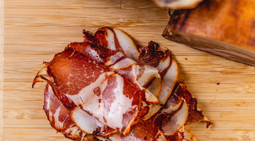 Two chubs of coppa and next to thinly sliced coppa on a cutting board and picture from above.
