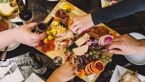 A picture shot from above of hands reaching to grab food from a charcuterie board full of meat, cheese, vegetables and fruit.
