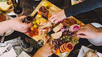 A picture shot from above of hands reaching to grab food from a charcuterie board full of meat, cheese, vegetables and fruit.