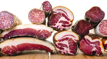 A picture looking at the cross section of several types of cured meat like salami, coppa, speck, spanish lomo and more. All the meat is stacked on top of each other and top of cutting boards.