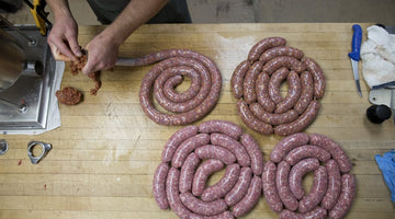 A picture of several sausages on a table while more sausage is being made with a casing stuffer.
