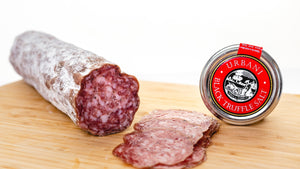 A chub of il porcellino salumi's large format Black Truffle Salami on the left side of a cutting board with thin slices in the foreground and a tin of black truffle salt to the right.