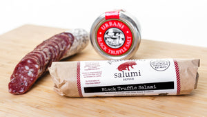 Two chubs of il porcellino salumi's retail sized black truffle salami on a cutting board. One chub is cut into slices and the other chub is in packaging with black truffle salt behind it.