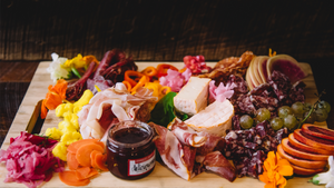 A darkly lit charcuterie board displayed various cured meats, cheeses, fruits, vegetables and a jar of jam.
