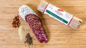 Two chubs of il porcellino salumi's Cacciatore salami on a cutting board and pictured from above. One salami chub is in packaging and the other one is cut into slices with herbs and spices next to it.