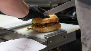 A picture of a sandwich on a preparation table with a knife cutting into it and a hand steadying the sandwich.