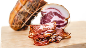 A picture of il porcellino salumi's Coppa cut into slices and whole muscle Coppa chubs in the background. The Charcuterie Masters grand champion seal and Good Food Awards 2017 winner seal are on the picture.