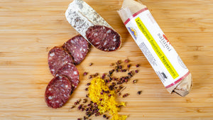 Two chubs of il porcellino salumi's Lemongrass Szechuan Peppercorn salami pictured from above on a cutting board. Half of one chub is cut into slices and the other is in packaging with herbs and spices between them.