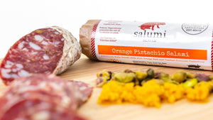 Two chubs of il porcellino salumi's orange pistachio salami pictured from the side on a cutting board. One of the chubs is cut into slices and the other is in packaging with herbs and spices in the foreground.