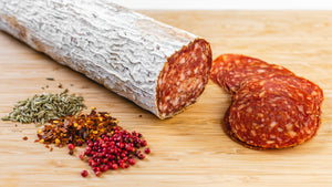 A chub of il porcellino salumi's large format Soppressata Salami pictured at angle from above with Soppressatta slices, herbs and spices next to it.