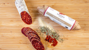 Two chubs of il porcellino salumi's Spanish Chorizo salami pictured from above on a cutting board. One of the chubs is cut into slices and the other is in packaging with herbs and spices between them.