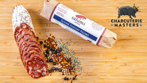 Two chubs of il porcellino salumi's Spiced Juniper salami pictured from above on a cutting board. One of the chubs is cut into slices & the other is packaging with herbs & spices between them. The charcuterie masters grand champion seal is on the image.