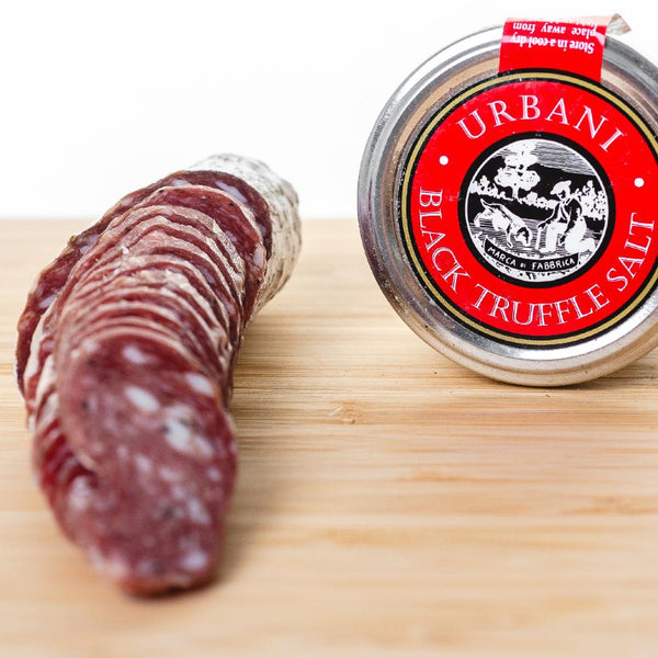 An up close picture of black truffle salami cut into slices on a cutting board next to a jar of black truffle salt.