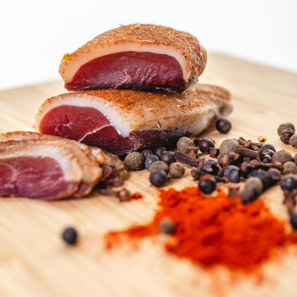 Chunks of duck prosciutto on a cutting board with herbs and spices in the foreground.