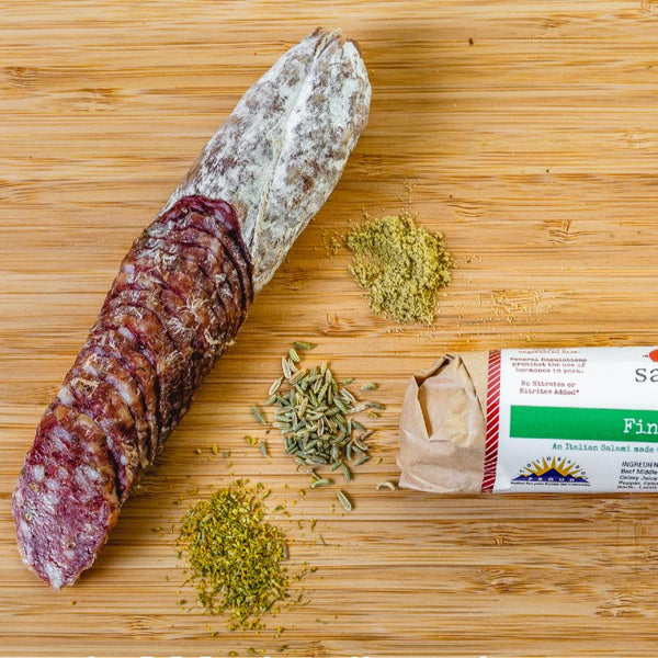A product photo of il porcellino salumi's Finocchiona salami shot from above. Half of the salami in packaging is cropped out of the image and another chub of salami is cut into slices.