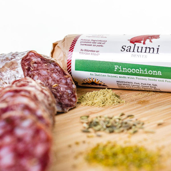 A close up picture of Finocchiona salami on a cutting board with herbs and spices. One salami is in packaging and the other is cut into slices.