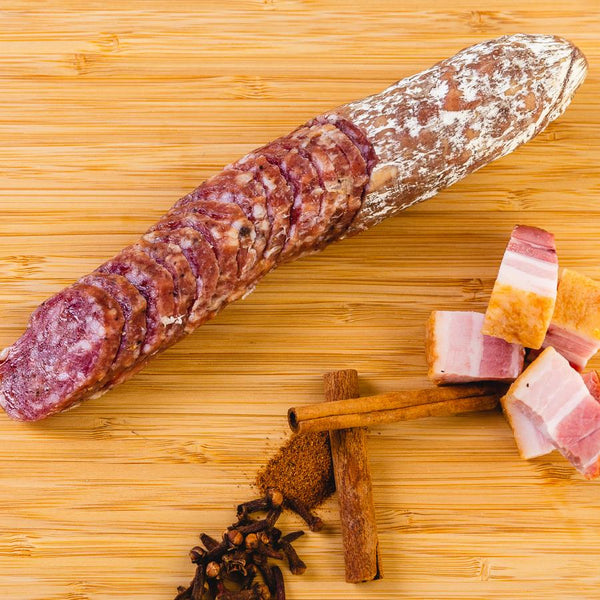 A picture of bacon whiskey salami shot from above. The salami is cut into slices in a cutting board next to herbs, spices and chunks of bacon.