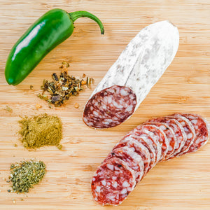 A product photo of green chile tequila salami shot from above. Half of the salami is whole and the other half is cut into slices on a cutting board with a green chile, herbs and spices.