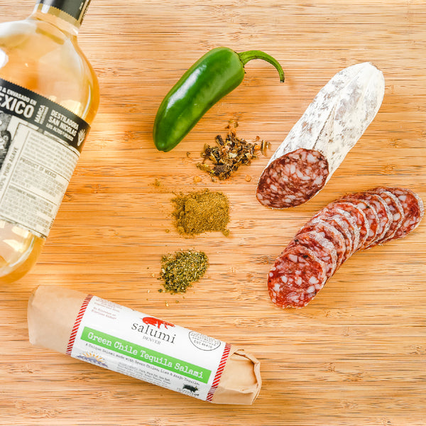 A product photo of green chile tequila salami shot from above. Half of the salami is whole and the other half is cut into slices on a cutting board with a tequila bottle, a green chile, herbs and spices.