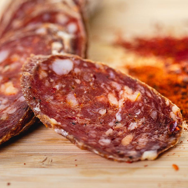 A close up of salami slices displaying the fat marbling, herbs and spices. The salami slices are on a wood cutting board with blurred herbs and spices in the background.