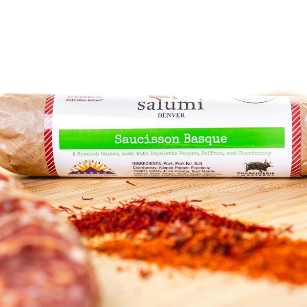 A close up of Saucisson Basque salami in packaging on a cutting board with blurred salami slices, herbs and spices in the foreground. 