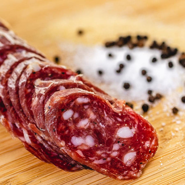 A close up of salami slices displaying the fat marbling, herbs and spices. The salami slices are on a wood cutting board with herbs and spices blurred in the background.