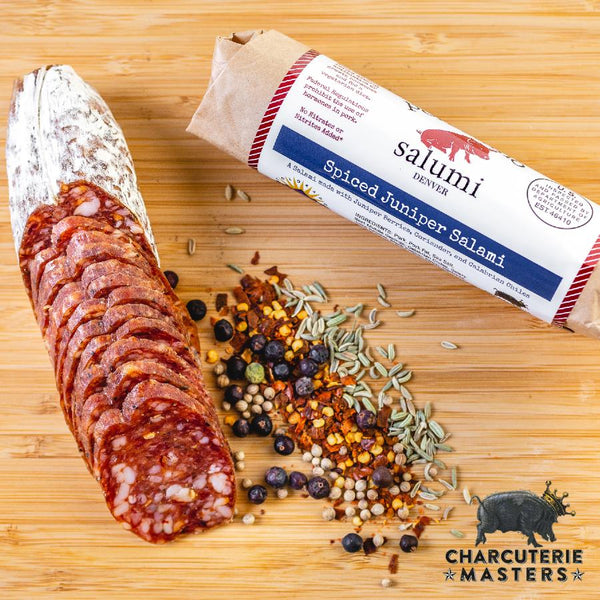 A picture of il porcellino salumi's Spiced Juniper Salami on a cutting board with herbs and spices shot from above. Once piece of salami is cut into slices while the other remains in packaging. Plus the Charcuterie Masters Grand Champion logo is placed in the lower right corner of the image.