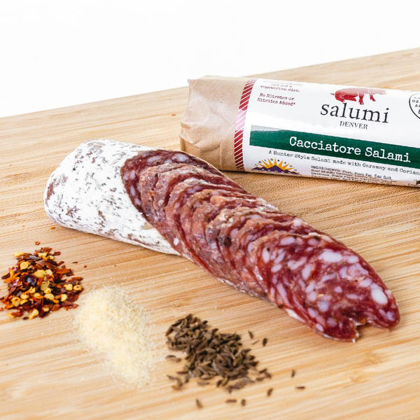 Cacciatore salami in packaging and cut into slices on a cutting board next to herbs and spices.