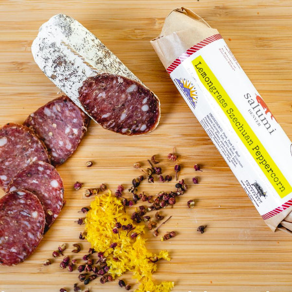 A product photo of il porcellino salumi's Lemongrass Szechuan Peppercorn Salami shot from above. A chub of salami is on a cutting board next to salami slices, packaged salami, herbs and spices.